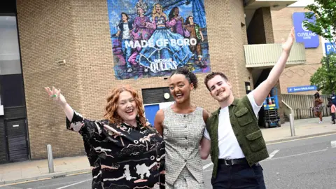 Actors Patsy Lowe, Alexandra Mardell and Elijah Young stand with their arms stretched out and smile. The mural is in the background on a the outside of a large brick building. It features the cast next to the Transporter Bridge and says Made of Boro.