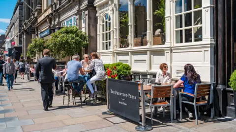 People sitting at tables eating and drinking on the street in the sunshine.