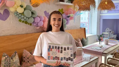 Amelia Lord Amelia Lord holding a book or folder open with the numbers 2026 across it and images of books she has read or wants to read in that year. She has long straight hair held back by a headband, wears a white tshirt and jeans and is smiling at the camera. A bench with soft patterned cushions is behind her with two tables draped with soft pink cloth and vases of flowers. There is a balloon display on the walls in pastel colours.