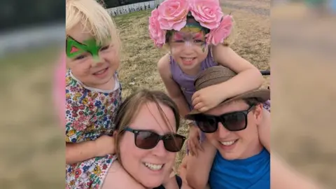 Family handout Vicky with her husband, both smiling and wearing sunglasses, with their two daughters sat on their shoulders. The two girls have face paint on and one has pink flowers in her hair.