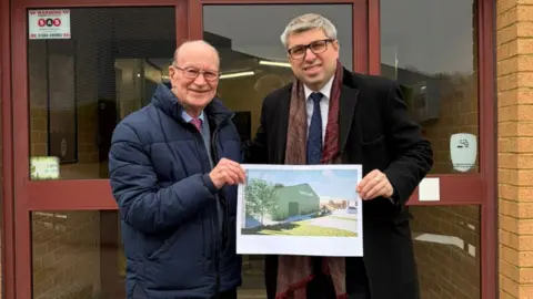 Two men standing outside a set of double doors hold up an poster showing the design of a proposed new sports facility.