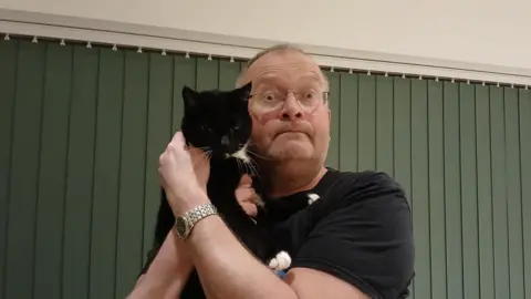 Ronan Munro Ronan has short grey hair and wears small glasses and a black t-shirt. He is holding up a black and white cat.