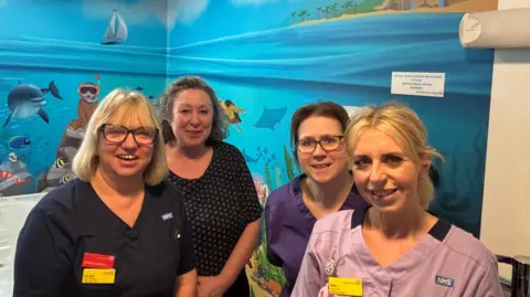 Four women standing in a hospital ward bathroom. Its walls are covered in a colourful mural which shows the sea, a beach, fish, dolphin, animals snorkelling and plants. 