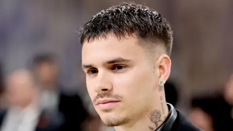 Getty Images A head and shoulders shot of Romeo shows him at the Met Gala wearing a black jacket and shiny black shirt. He looks expressionless as he stares to the left of the image, with other guests out of focus over his right shoulder.