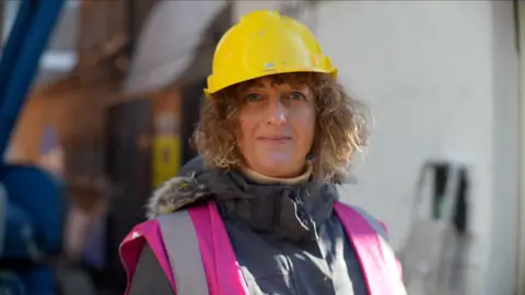 Jamie Niblock/BBC Iona Hodgson smiles at the camera. She has short curly light brown hair that can be seen underneath a yellow hard helmet she is wearing. She also wears a pink hi-vis jacket above a grey coat.