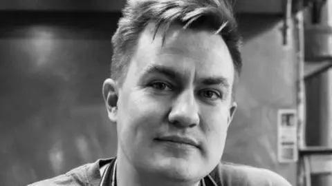 Adam Armstrong A close-up black-and-white image of Adam Armstrong, in a kitchen. He has short hair and is smiling.