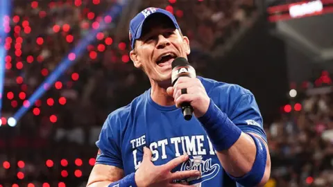 John Cena, a man wearing a blue cap, t-shirt and sweat bands, speaking into a black microphone at WWE's Raw event in Los Angeles