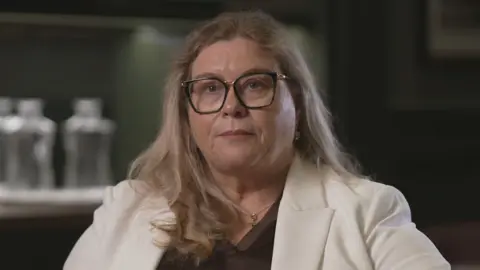 A woman, wearing a cream jacket and brown blouse, is looking at the camera. She has long blonde hair which hangs over her shoulders and is wearing large, dark-framed glasses. Behind the woman is a dimly lit room which is out of focus. Some large glass jars can be seen at one side under a spotlight. 