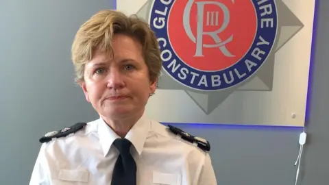 Carmelo Garcia Maggie Blyth, who has short light brown hair, looking straight ahead. She is wearing her uniform of a white shirt and black tie with black patches on her shoulders. The logo for Gloucestershire Constabulary is behind her on the wall.