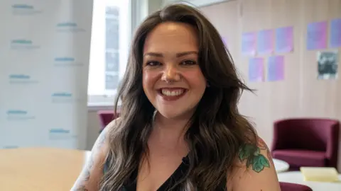 A photo of Lauren Burke looking straight at the camera. She is indoors in some kind of office. She has long brown hair.