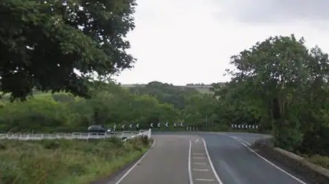 Google The area of the Blackboards, which is a sharp bend with a junction joining from the right. There are black-and-white chevrons on the edge of the bend and trees on either side.