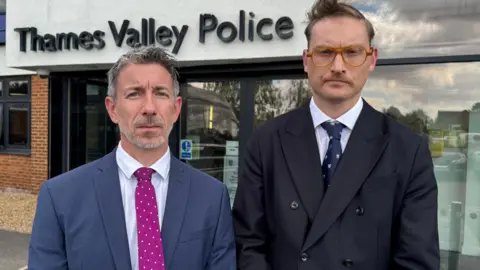Two men in suits - one slightly shorter than the other - looking into the camera with serious facial expressions. They are stood in front of Thames Valley Police's Headquarters.