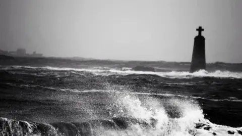 Aurora Craig Waves crash violently in a stormy sea, with a tall stone monument topped by a cross rising in the distance.