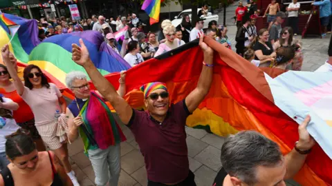 A group of people standing gin a line which holding a giant Pride flat. There is a man in the centre of the photo wearing a rainbow bandana and dark, rectangular sunglasses. He is holding his arms in the air and smiling into the camera. There is a woman behind him with white, short hair and glasses and she is wearing a priest's white collar with a black sleeveless shirt. Behind her is a woman with brown hair and sunglasses who is holding her arm up to the camera. There are lots more people walking around them.