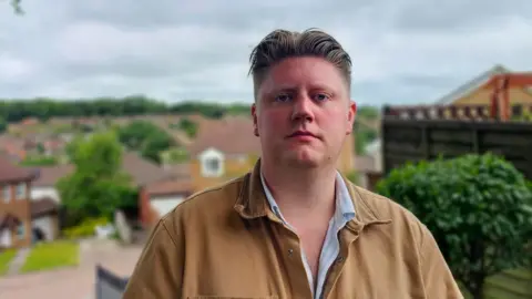 Councillor Robbie Lammas stands with his council ward in the distance behind him. In the foreground Councillor Lammas is wearing a beige jacket and looking towards the camera 
