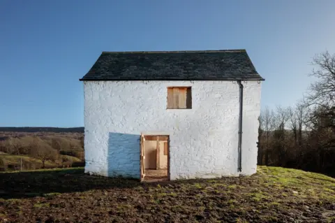 Historic England Henry's Castle. It is a small white building with a pitched roof set in the middle of green countryside. It has a small widow and an open door with some wooden features visible inside.