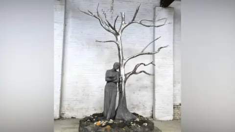 A statue including a shiny metal tree without leaves and a person leaning up against it on a scenic base with a white brick wall behind it