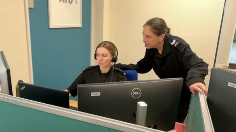 BBC TVP PC Harley Edwards and Sgt Dom Lucas from the RVR team in front of a computer. PC Edwards has a headset on and appears to be on a call. Sgt Lucal is over her shoulder.