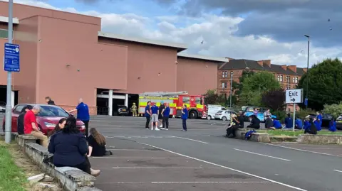 People gathered in a supermarket car park while a fire engine is parked beside a large retail building. Several individuals sit on kerbs and grassy areas, with others standing nearby. Cars are parked around the site under a cloudy sky, and an exit sign is visible in the foreground.