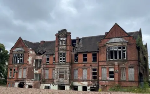 City of Wolverhampton Council A derelict former hospital building in red brick with lighter coloured stone detail including first-floor bay windows and a plaque reading Eye Infirmary. Ground flood windows have been boarded up, and parts of some walls demolished. The roof has gaping holes in the tiles.