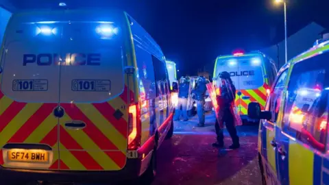 Three police vans, with police officers holding riot shield standing in the middle