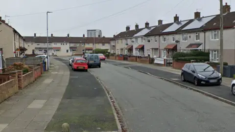 A street view of Madryn Avenue. The street is lined with houses and cars parked on the pavements.