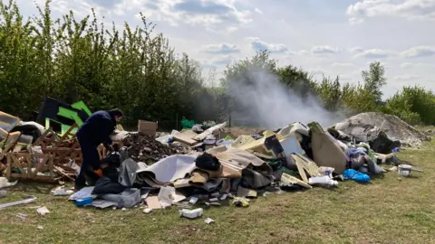 Gravesham Borough Council An investigator searches through piles of dumped waste, including carpets, furniture, timber and household rubbish, while smoke rises from a heap in the background.