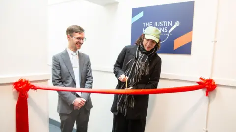 Justin Hawkins has long curly brown hair and is wearing a green cap, black and white scarf, black coat and trousers. He is cutting a red ribbon with gold scissors. A man stands next to him with brown hair, glasses and a grey suit with a white shirt. On the wall behind Justin is a navy blue sign that reads "the justin hawkins recording studio"