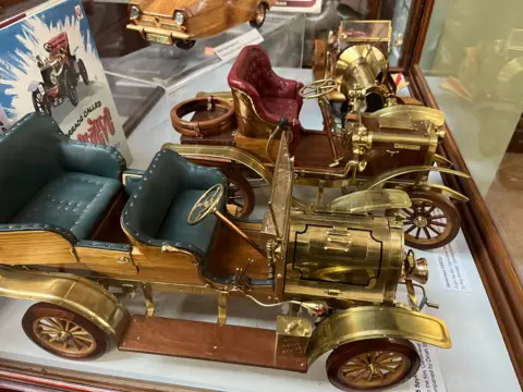 John Devine Four model cars in a display case, some are brass and some wood, the Chitty Chitty Bang Bang vehicle is at the back on the right with other ornate late 1800's cars at the front with open tops and leather seating, all made by Mr Clarke.