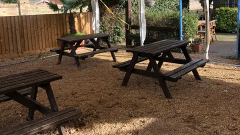 Bev Rimmer/BBC Three dark brown picnic benches stand on a square of gravel, underneath the canopy of a white marquee. A tan-coloured fence is to the left of the picture. The sun is shining. There are some green hedges in the background.