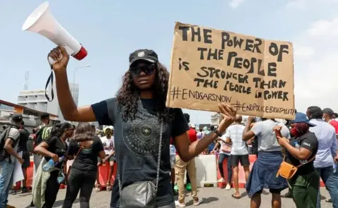 Reuters A woman in a black T-shirt, cap and sun glasses holds a loud hailer on her right hand and a cardboard sign in her left. The sign reads: "The power of the people is stronger that the people in power. #EndSars, ~EndPoliceBrtuality"