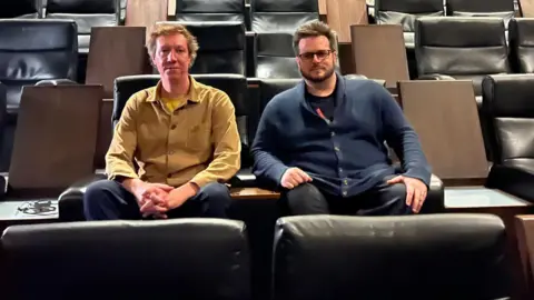 Jon Foster and James Lamont sit next to one another in a cinema, which has leather seats and wooden tables. Jon has longer greying hair, and is wearing a camel coloured shirt, as he clasps his hands together between his spread legs. James is wearing a navy cardigan and black rectangular glasses, and is sat with his hands resting on his knees. 