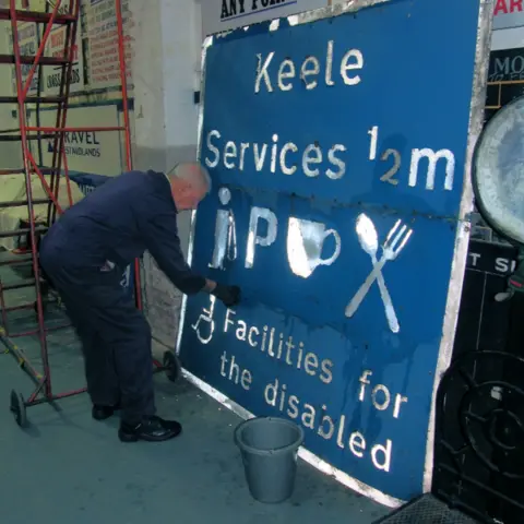 National Highways The Keele Services road sign, which is blue with white writing and a white border, which is being painted on by a man with silver hair and a blue boiler suit, who is bending down to paint it