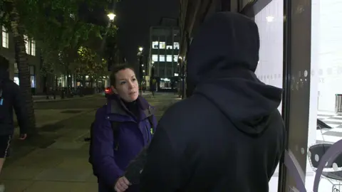 Journalist Frankie McCamley in a purple jacket interviewing a person in a dark hooded sweatshirt on a city street at night. The journalist is facing the person, who has their back to the camera.