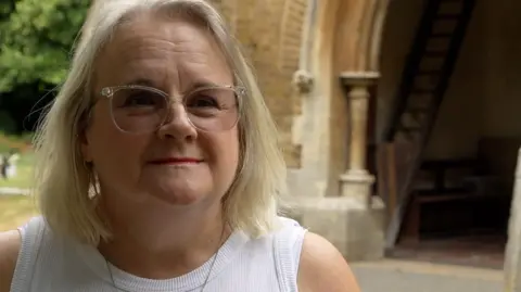 A woman wearing a white vest top with a crucifix necklace on. She has blonde hair and is standing at the entrance of a church.