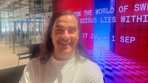 Robin McNicholas stood outside the Aviva Studios venue in front of a large colourful sign showing the date of the exhibition