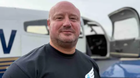 a white man with a bald head, wearing a black t-shirt with a wing logo on it, smiles at the camera in frnt of a blurred out aircraft.