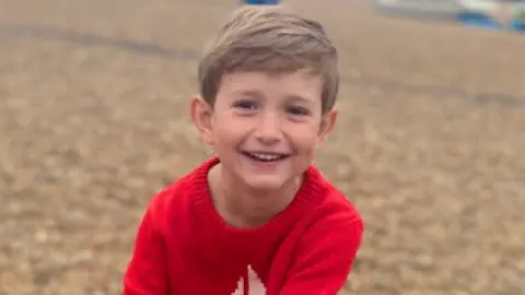 A boy with a red knitted jumper with a white boat on it. He is smiling and has blonde hair. In the background is a beach with beige pebbles