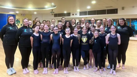 A photo of a dance group standing in a formation smiling into the camera. There are around 25 children in the photo and 3 teachers. Everyone is wearing black. They are in a bright studio with wooden floors. 