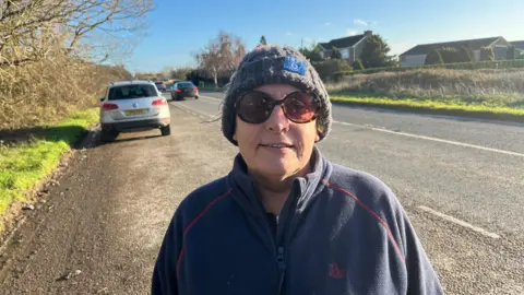 John Devine/BBC Alison Searle, 58, has a grey woollen bobble hat on with a blue square patch on the front, she is wearing large dark glasses and has a blue fleece top on, the A605 road is behind her and some housing is visible in the distance, a car is in motion on the road and another is in a layby on the left.