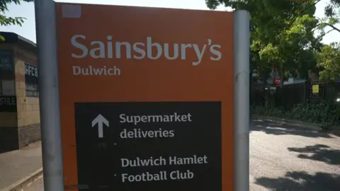 An orange and black road sign reading "Sainsbury's Dulwich" with an arrow pointing straight ahead for "Supermarket deliveries" and "Dulwich Hamlet Football Club".