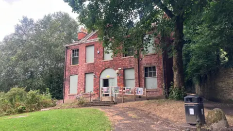 Photograph of Rock Hall in the Moses Gate Country Park in Farnworth. The image shows boarded-up windows and the building is surrounded by trees.