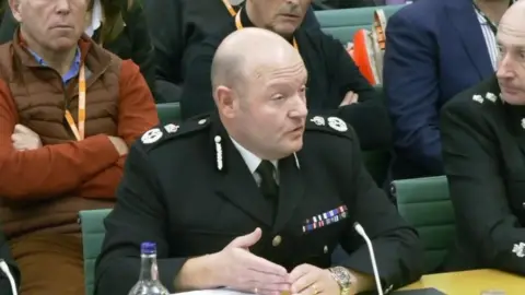 A man wearing a black police uniform sits in front of MPs. Rows of people can be seen behind him.