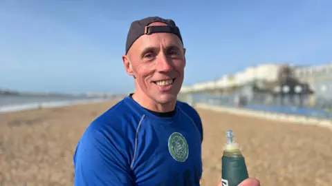 George Carden/BBC Darren Evans standing on Brighton beach wearing a blue "The Green Runners" top smiling at the camera