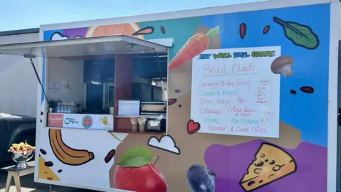 Caring Cooks of Jersey "Spud Truck" pictured which is a trailer that has been painted with a bold pattern including blue, purple, brown, white and green. Cartoon-looking fruit and vegetables are also painted on the truck. 