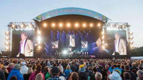 PA A large crowd of people can be seen from behind, looking upon a massive stage. At the top of the stage, in pink lettering, are the words "Isle of Wight Festival", on a colourful blue arch with rainbows and other bright patterns. On the stage we can just about make out the figure of Rod Stewart. The singer's image is amplified on large screens either side of the stage. He's wearing a white collared shirt under a shiny gold jacket and appears to be in good spirits. 