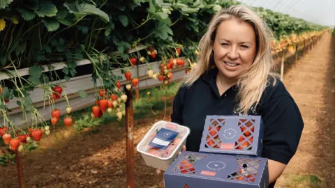 Annabel's Deliciously British A woman with long blonde hair wearing a black polo shirt holds punnets of strawberries on a farm