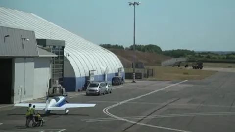 A hangar is on the left and has several vehicles and a small plane outside of it. Next to it is part of the runway slip at Cambridge Airport, which has small vehicles on it.