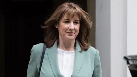 Rachel Reeves walks out of Number 10 Downing Street wearing a white blouse and light blue jacket smiling slightly with her mouth closed