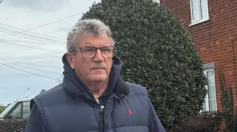 Terry Penton has grey hair and is wearing glasses, a navy bodywarmer and hoodie. He looks serious and is standing outside a brick house.
